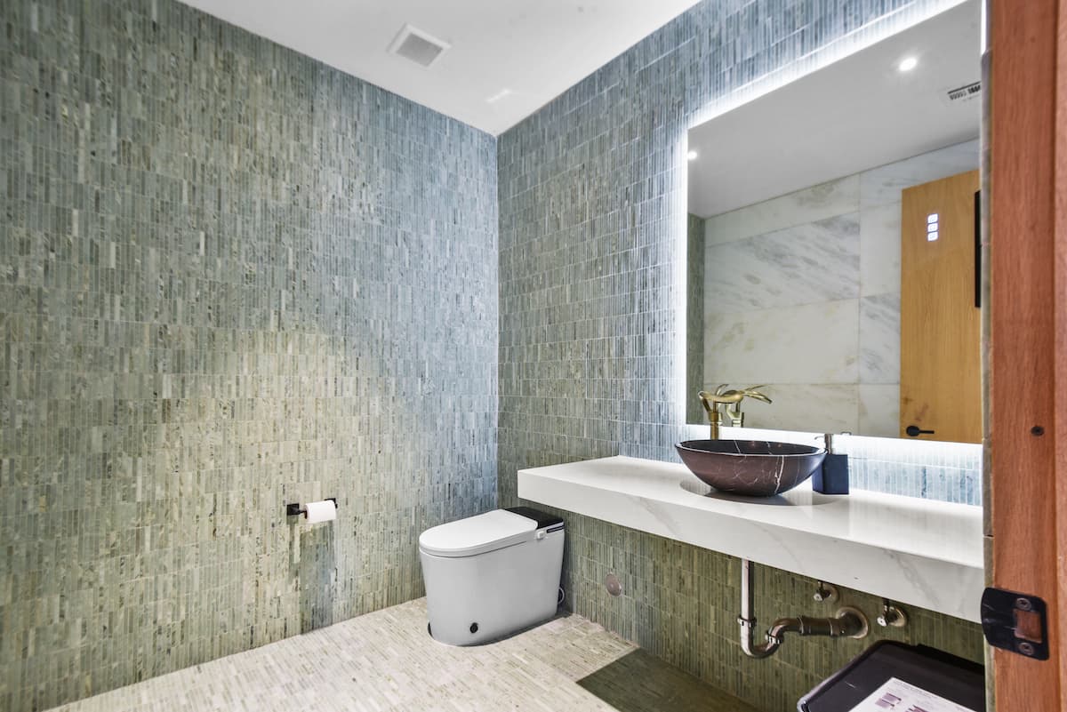 Restroom with green mosaic tile walls, a backlit mirror, marble counter and stone vessel sink